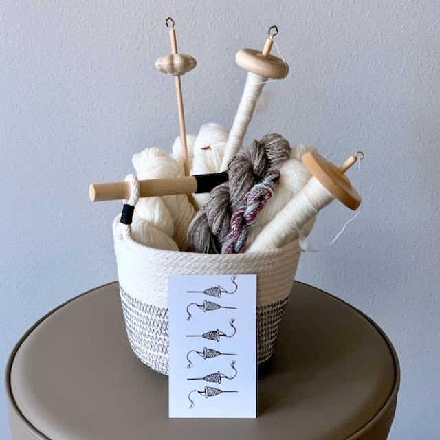 A rope coil basket filled with drop spindles and skeins of handspun yarn. In front of the basket is an index card stamped with a repeated drop spindle image.