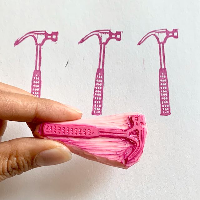 Fingers holding a pink carved stamp of a hammer, held above 3 pink block prints created from the block.