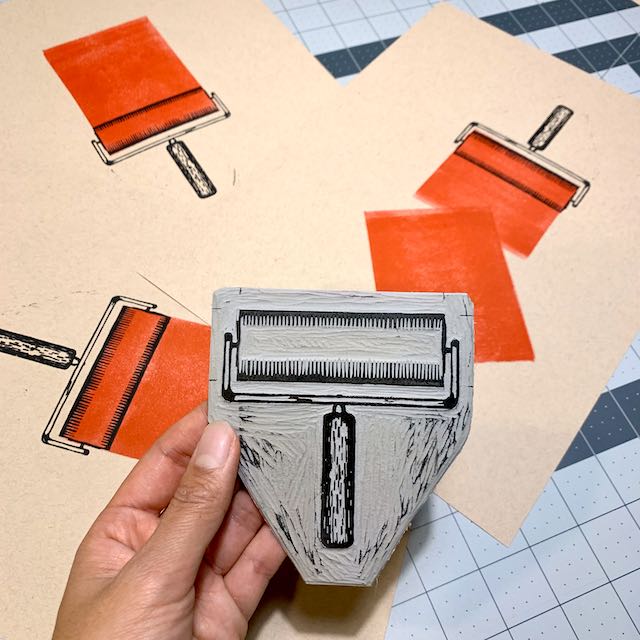 A hand holding a carved linoleum block of a brayer, in front of several paper prints of the same design.