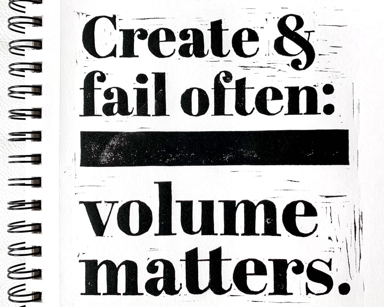 A blockprint in black ink on a white sketchbook, proclaiming in large serif font: Create and fail often - volume matters.