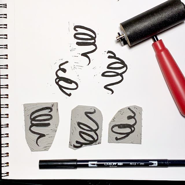 Three carved linoleum blocks depicting black squiggles, impressions from the 3 blocks, a black brush-tip pen, and a red-handled brayer, all sitting on a white spiral-bound notebook page.