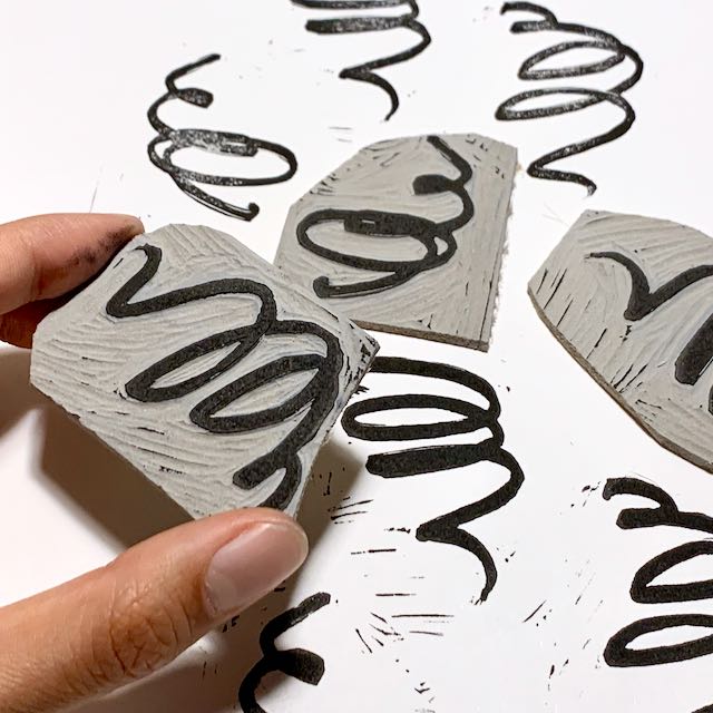 Fingers hold up a carved linoleum block of black squiggles. Two similar linoleum blocks, as well as relief prints from all three, lie on the notebook in the background.