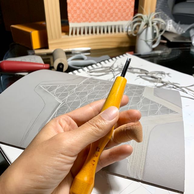 A hand holding a carving tool above a partially carved linoleum block. The hand's ring finger is covered in a bandaid.