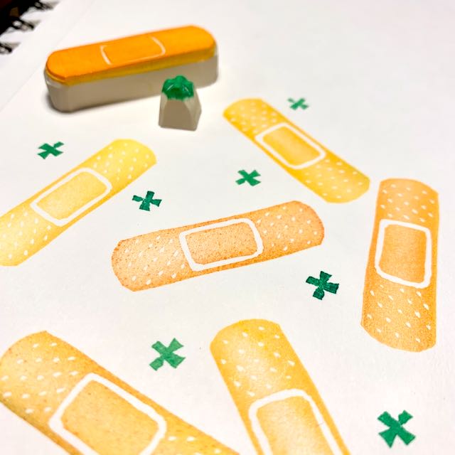 An array of printed orange bandaids and green health symbols. The two stamps used to create these prints sit in the background.
