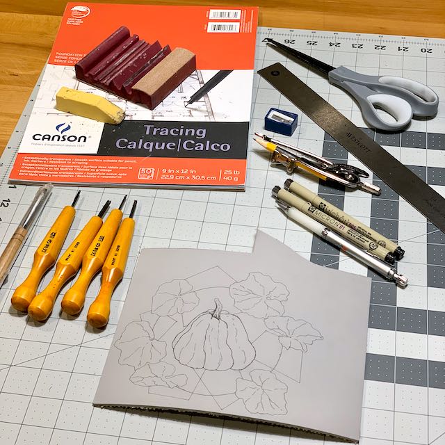 A worktable covered in tools, ready to carve a pumpkin design into a flat sheet of linoleum.