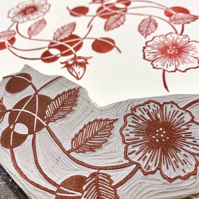 An up-close view of a carved linoleum block depicting a floral design, inked in dark red ink. The relief print from the block is visible in the background.