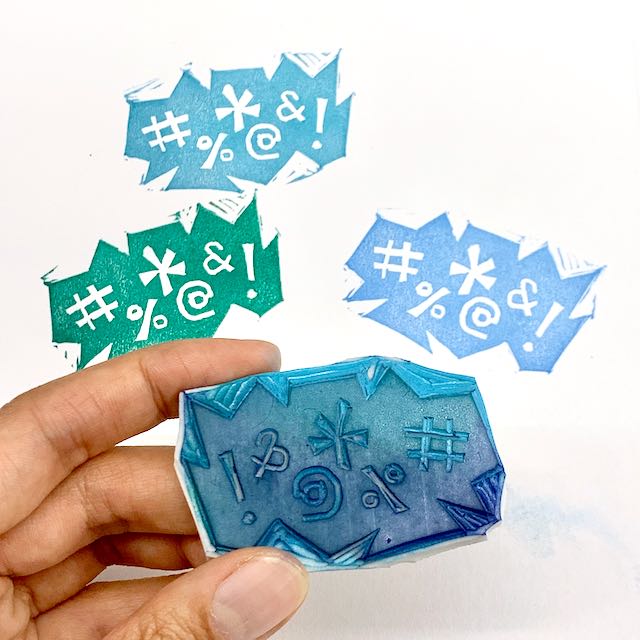 Fingers hold up a blue carved block of a grawlix. Three prints in different colors are on the paper in the background.