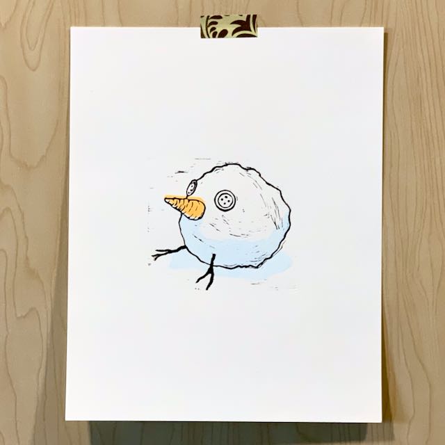 A print depicting a snowball with a carrot nose, 2 button eyes, and 2 stick legs. The print is on white paper and is taped to a wooden backdrop.