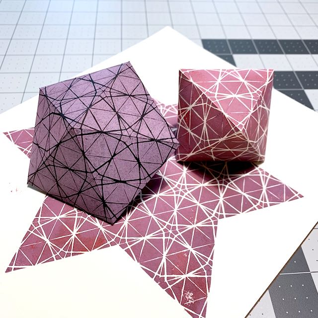 Two polyhedral shapes folded from linocut-printed paper. The left shape has 10 sides and is purple on black paper; the right shape has 8 sides and is pink-purple on white paper. Both are printed in a kumiko geometric pattern.