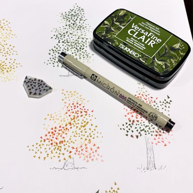 A flatlay of the tools used to create the print of trees in the background: a hand carved stamp of small triangles, a Micron pen, and a green VersaFine Clair stamp pad.