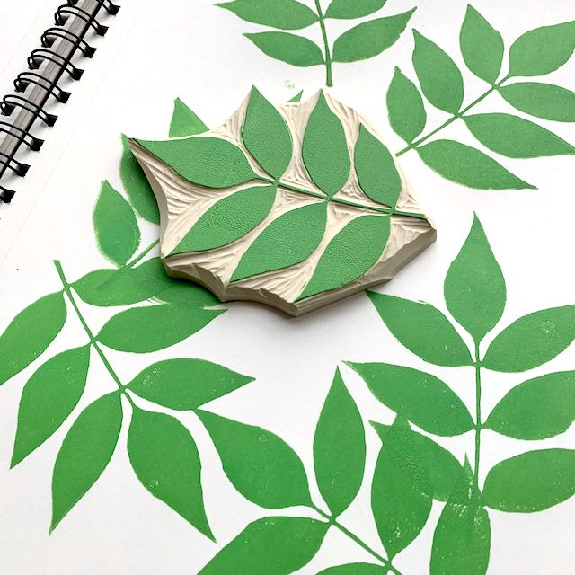 A carved block depicting an ash leaf, covered in green ink. The block sits on top of a white notebook covered in ash leaf prints.