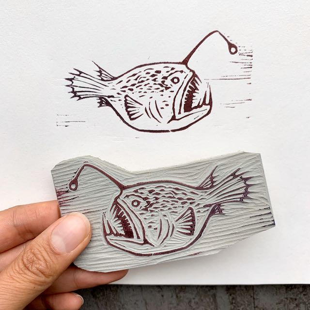 On top, a block print of an angler fish, facing to the right. On the bottom, a hand holding the linocut block used to create the print; the fish in the carving faces to the left.