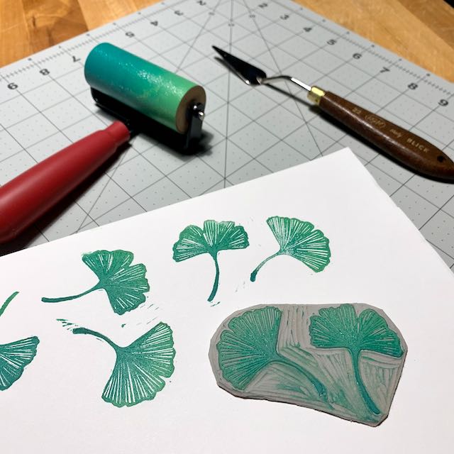 An array of ginkgo leaf prints, a carved linoleum block of 2 ginkgo leaves, and a red-handled brayer inked in a blue-to-green gradient.