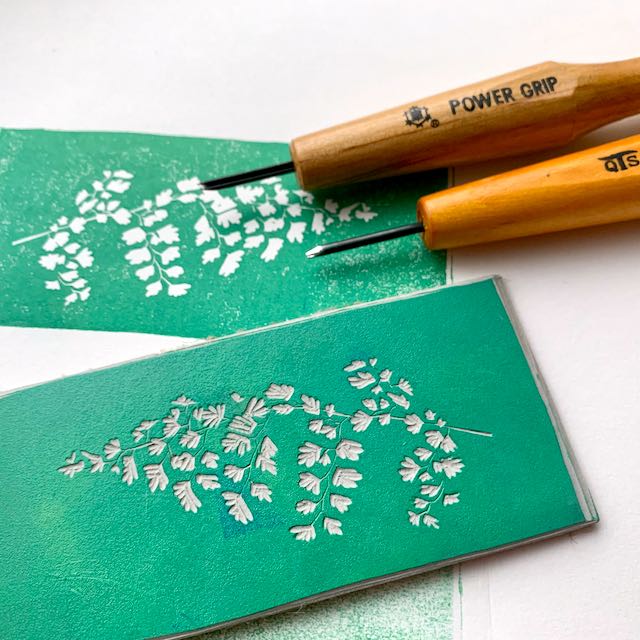 A carved linoleum block of a maindenhair fern, inked up in blue-green. In the background are 2 carving tools and a print created from the block, all on white paper.