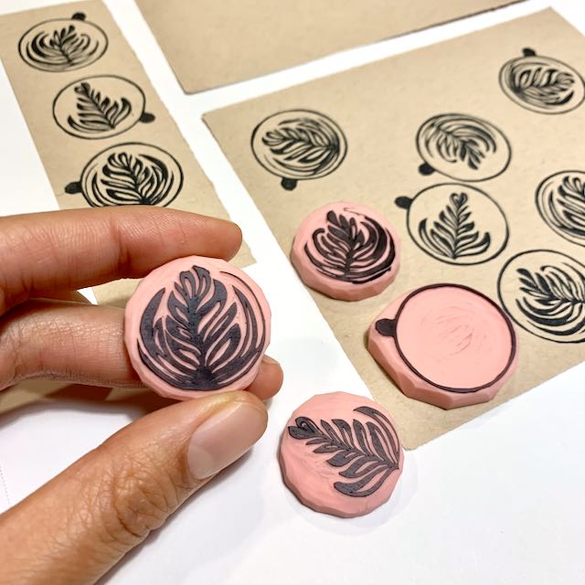 Fingers holding a tiny pink stamp of latte art. On the white backdrop behind the hand are 2 stamps of other latte art styles, 1 stamp of an empty coffee cup as viewed from above, and several sheets of brown paper covered in latte mug and latte art stamps.