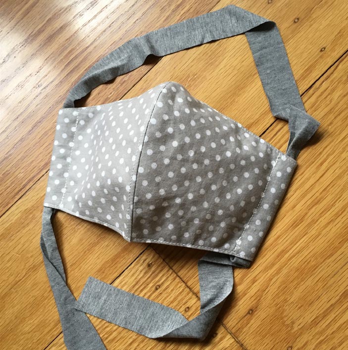 A grey and white polkadot fabric mask. The face mask is fitted, so that the center line rises above the edges in a gentle curve. A single grey fabric tie loops through channels at the side of the mask, and the loose ends hang from the bottom of the channels.