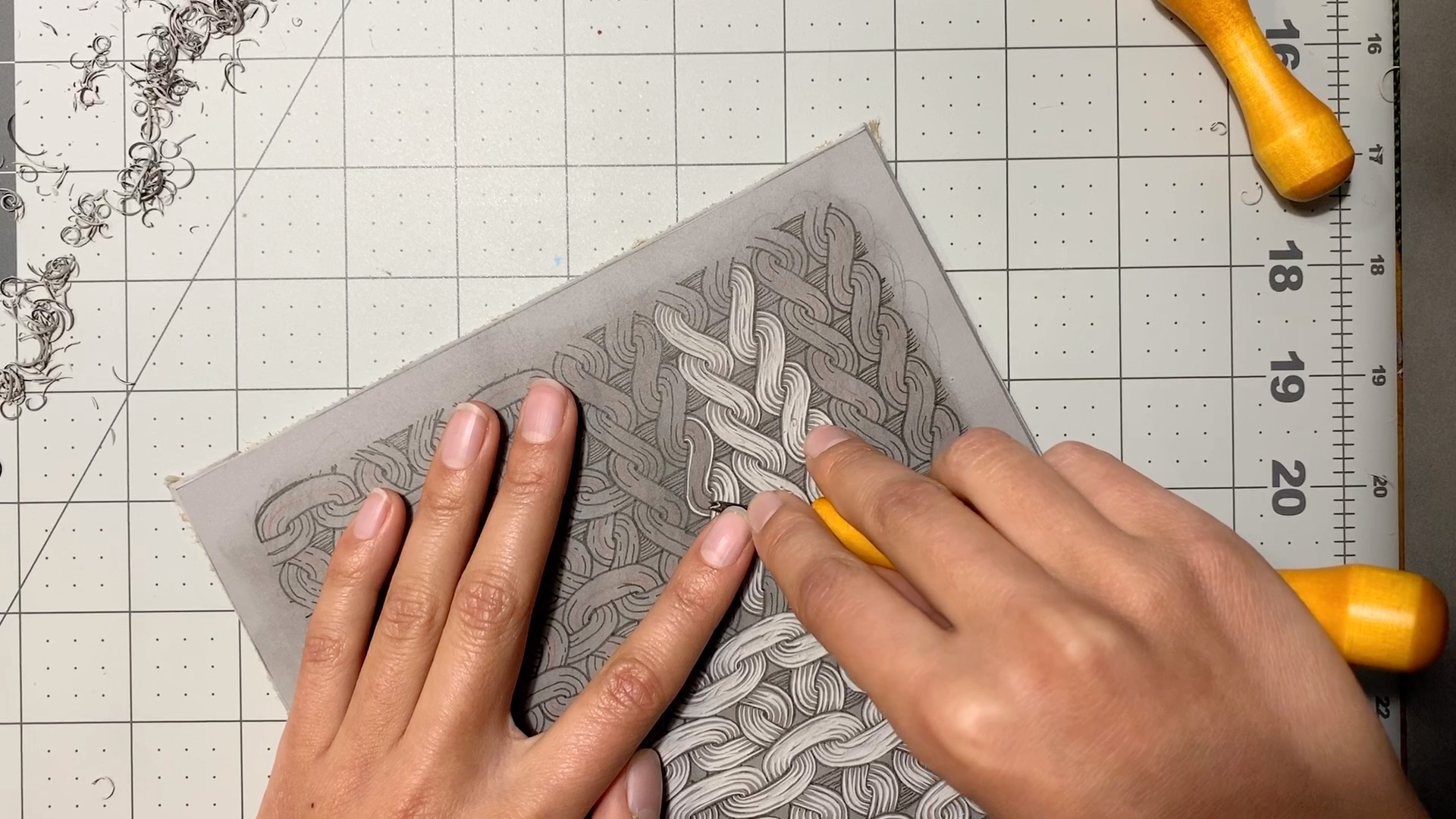 2 hands positioned on top of a grey piece of linoleum. The right hand holds a yellow carving tool and is removing material from the surface of the linoleum.