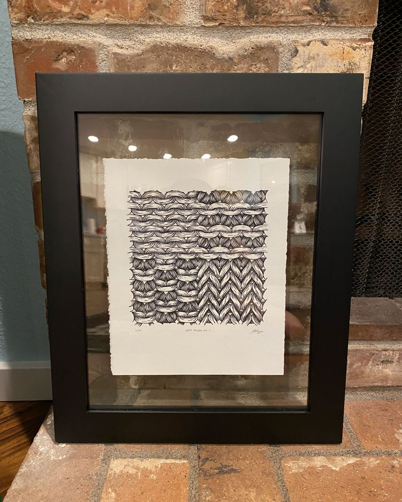 A print of knit stitch texture is sandwiched between two pieces of clear glass within a black frame. The frame sits on a brick fireplace.