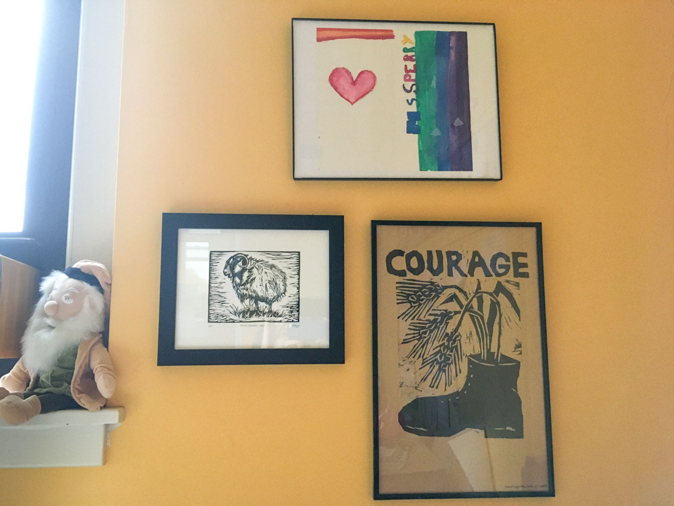 A sheep print within a black frame, hanging on a yellow wall next to two other pieces of artwork.