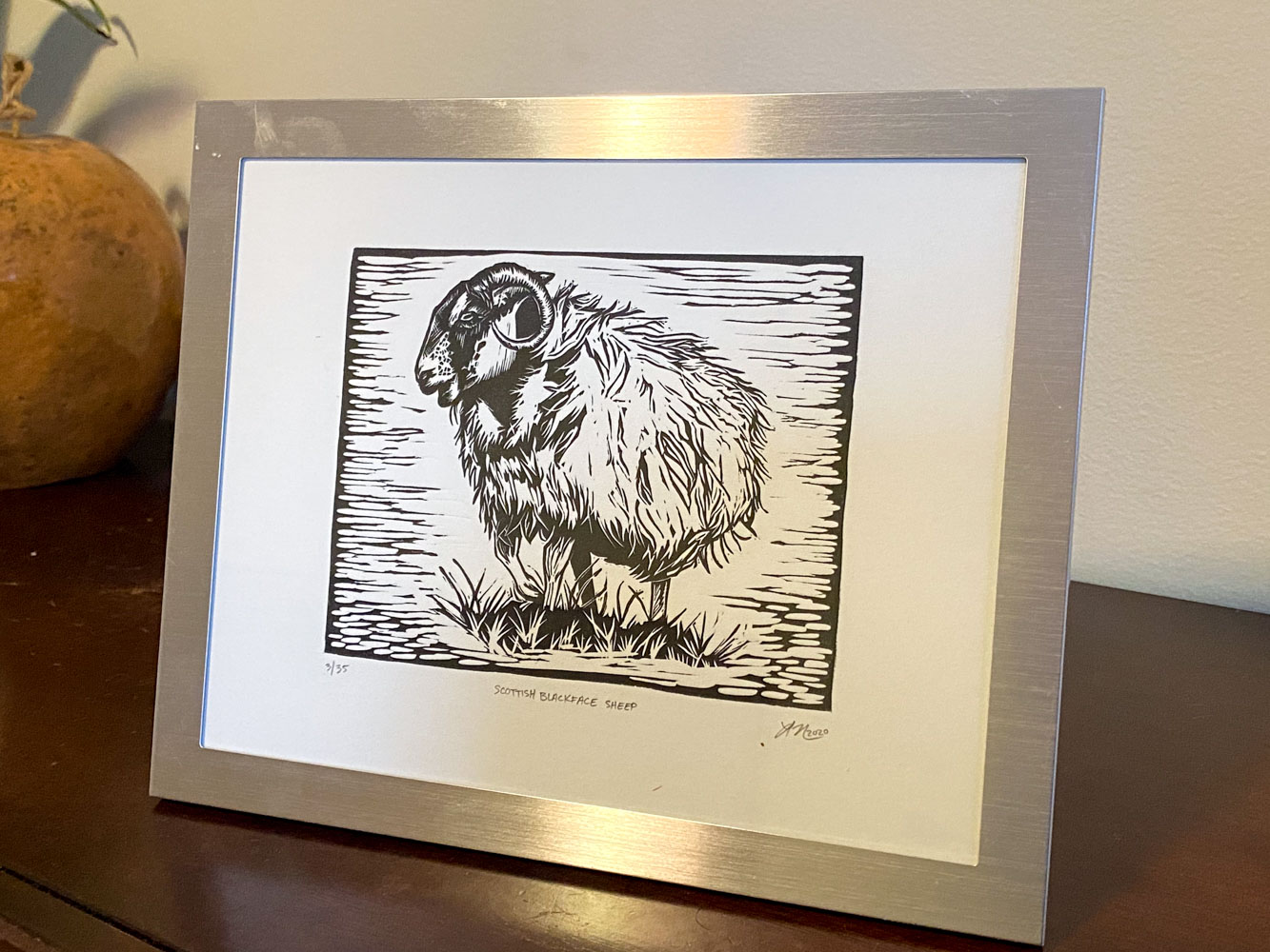 A sheep print displayed in a shiny metal frame. The frame is propped up on a wooden desk with a white wall in the background.