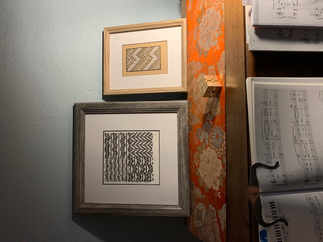 Two framed prints (the left of knit stitches, the right of twill weave structure) sit on top of an upright piano. Piano sheet music is visible at the bottom of the image.