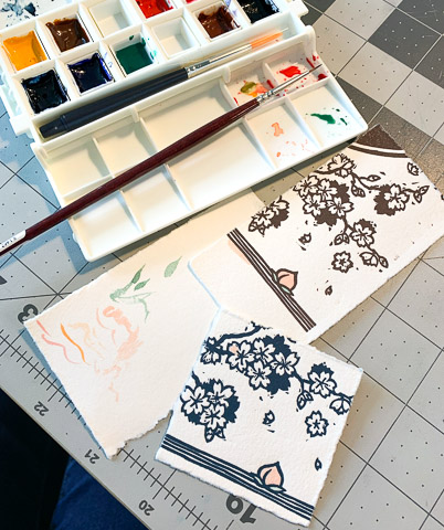 Two scraps of paper have been printed with a corner of the block, the bottom paper with blue and the top one with brown. A watercolor palette and watercolor brush sit next to the papers, and the peach visible in both scraps has been colored in pink and green.