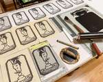 A table covered in horse blockprints, the linoleum block, a bamboo baren, and a glass inking plate covered in black ink and a brayer.