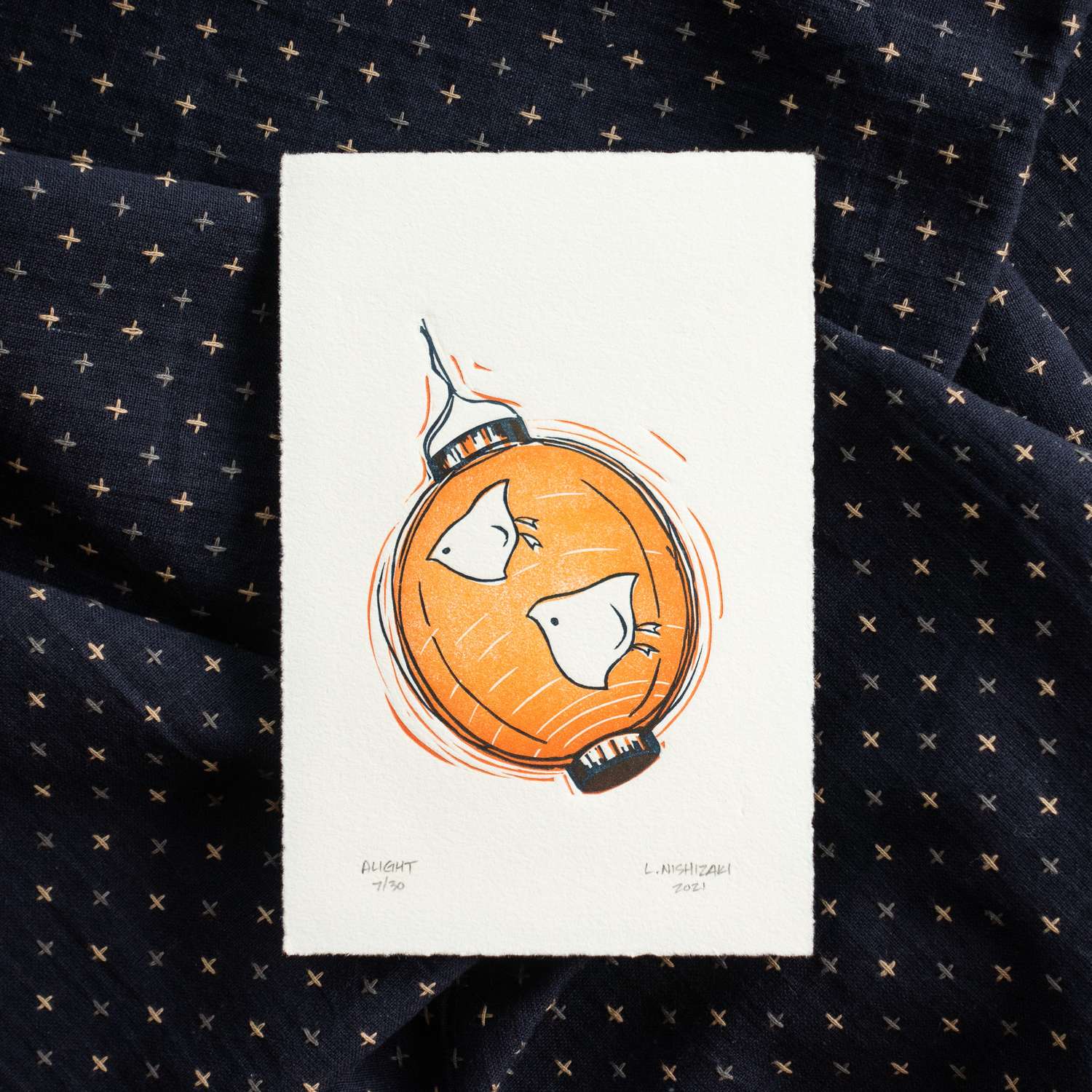 A rectangular print sits on top of crumpled dark navy fabric with light-colored checks. The print depicts an orange paper lantern with two chidori flying across the face towards the left. The lantern is drawn in sketchy lines to depict motion. The print is signed along the bottom edge.
