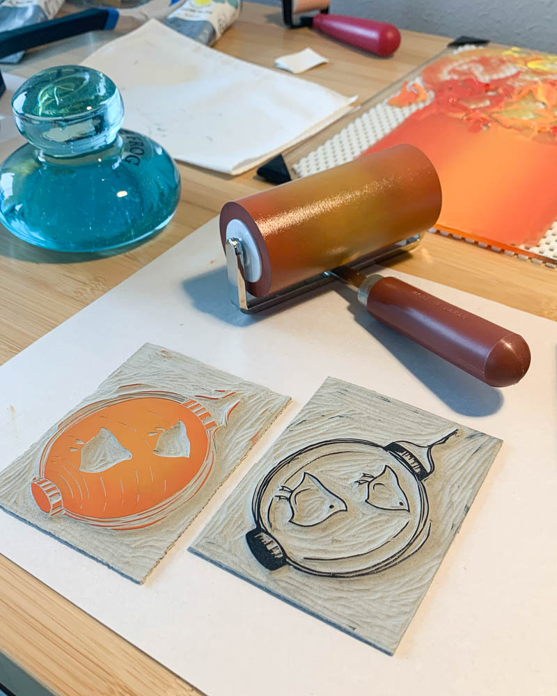 2 carved linoleum blocks inked in orange and navy, with a brayer, blue glass baren, and glass inking plate in the background