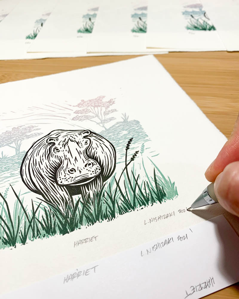 A hand signing the bottom of a paper print depicting a hippo
