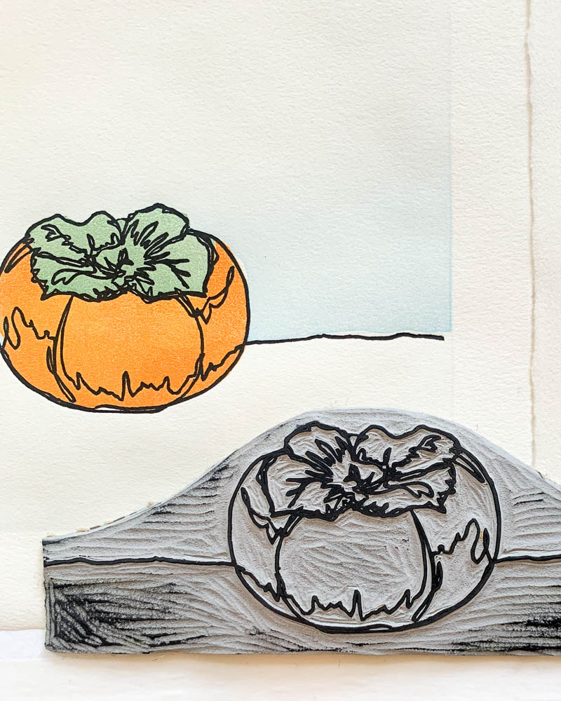 A carved grey linoleum block with a stylized persimmon, resting on top of a persimmon print