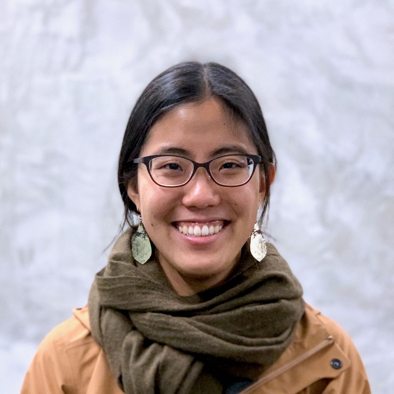 headshot of the artist, wearing a camel-colored jacket and brown scarf and standing in front of a grey wall