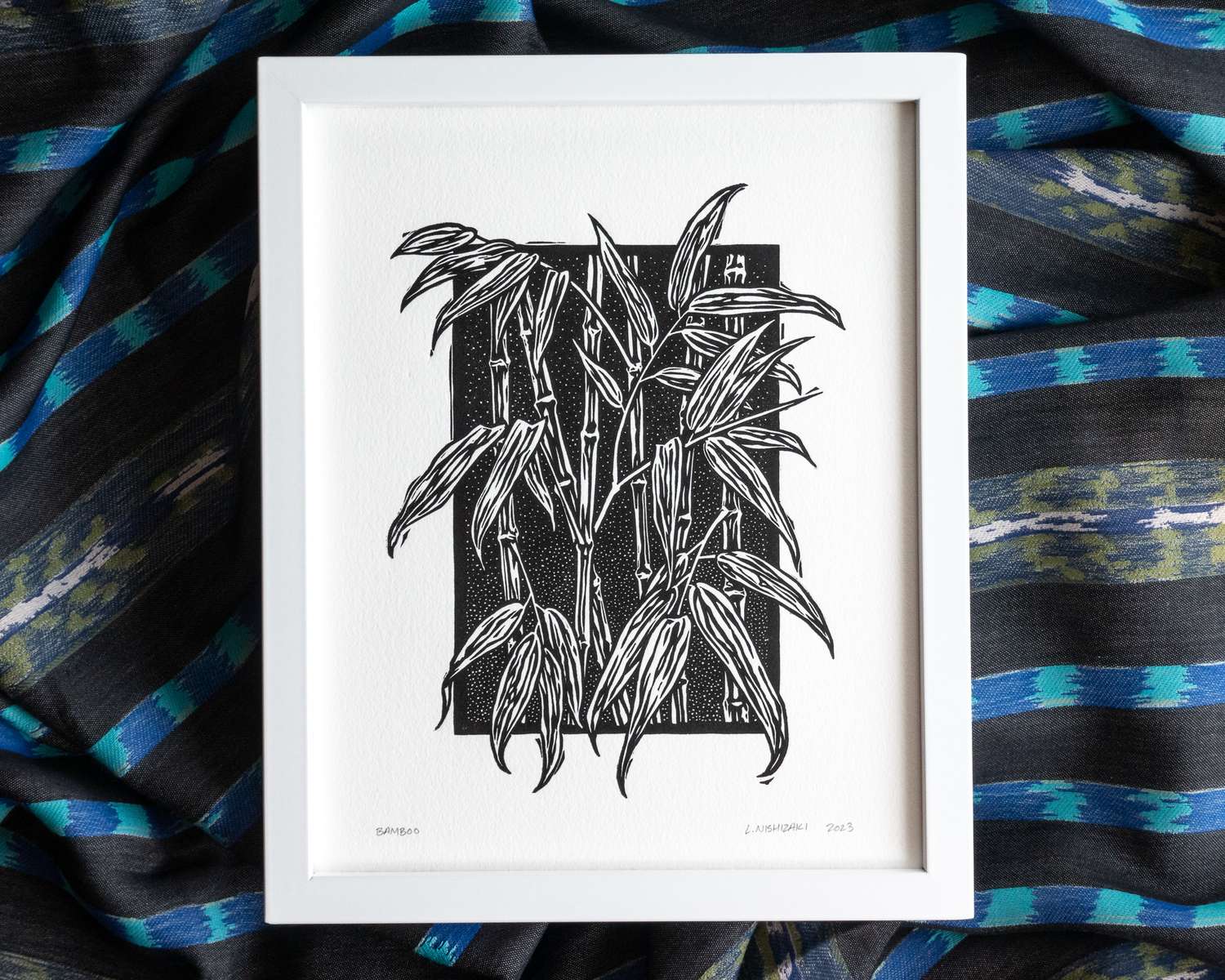A black and white blockprint of bamboo, printed on white paper and framed in a white wood frame. The frame sits on rumpled blue and black striped fabric.