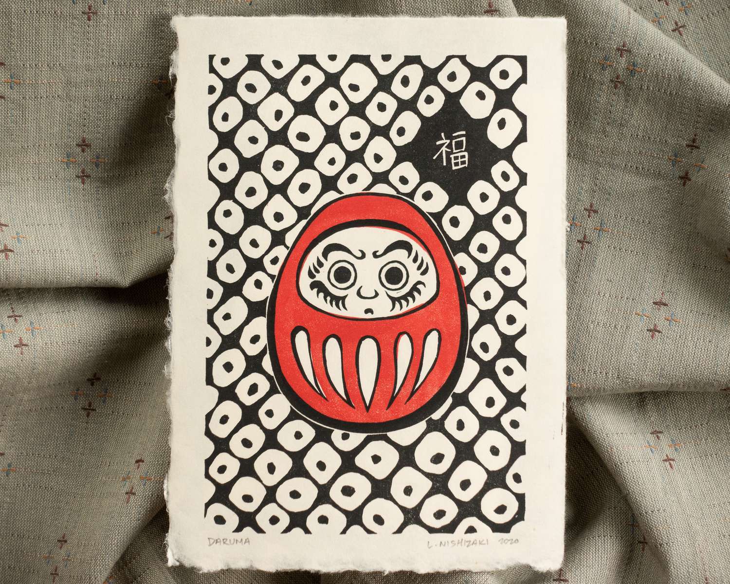 A rectangular print on light brown paper, depicting an egg-shaped Japanese daruma doll in red, and an all-over geometric background pattern. A Japanese kanji character meaning good fortune is in the top right.