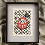 The daruma print lying on grey matboard inside of a black metal frame.