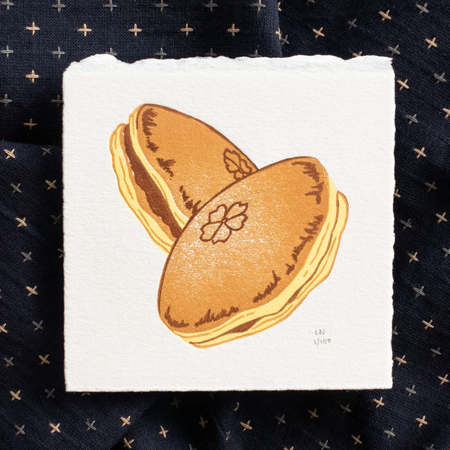 A square piece of paper depicting two overlapping dorayaki. Each dorayaki is printed in yellow, orange-brown, and dark brown, and are suspended in the air. The print sits on crumpled dark navy fabric with light-colored checks. The print is signed in the bottom right corner.