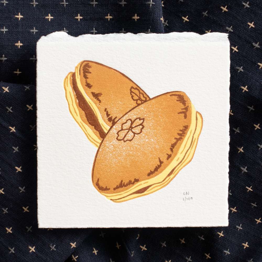 A square piece of paper depicting two overlapping dorayaki. Each dorayaki is printed in yellow, orange-brown, and dark brown, and are suspended in the air. The print sits on crumpled dark navy fabric with light-colored checks. The print is signed in the bottom right corner.