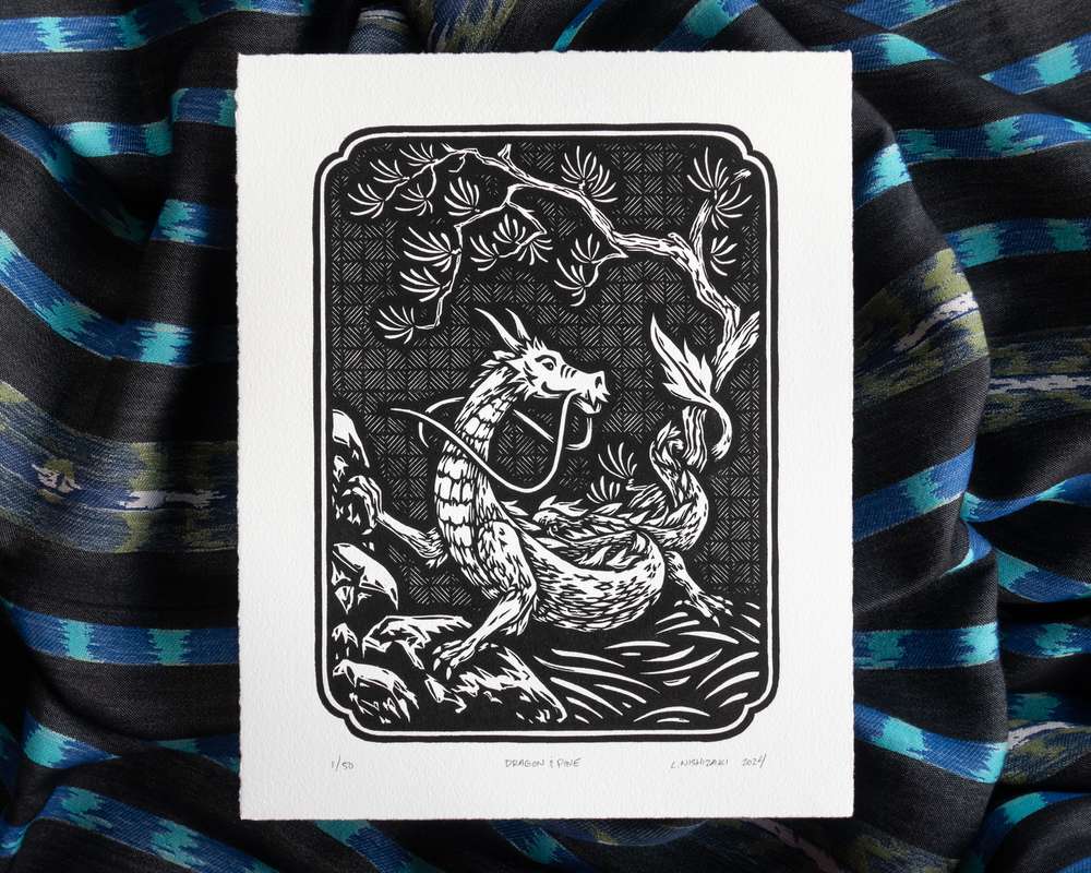 Black and white illustration of a dragon curled around rocks and a pine tree. The white paper sits on top of rumpled blue striped fabric.