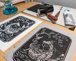 A messy desk covered with an inked linoleum block, glass inking surface, and brayer. A complete blockprint is in the background.