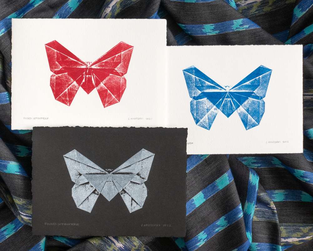 Three overlapping prints of butterflies on white paper, sitting on top of rumpled black and blue fabric. The butterfly at top left is magenta on white paper; top right is cyan on white paper; bottom left is white on black paper. All butterflies are in a geometric style, comprised of overlapping triangles printed with different ink density.