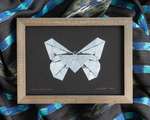 A white butterfly on black paper, within a brown-grey wood frame. The frame sits on top of rumpled black and blue fabric. The print is signed 'Folded Lepidoptera' in the bottom left corner, and 'L. Nishizaki 2022' in the bottom right corner.