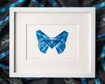 A blue butterfly printed on white paper, within a white mat, within a white frame. The frame sits on top of rumpled black and blue fabric. The print is signed 'Folded Lepidoptera' in the bottom left corner, and 'L. Nishizaki 2021' in the bottom right corner.