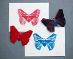 Two overlapping butterfly prints, one magenta and one blue, sitting on a grey backdrop. Two folded origami butterflies rest partly overlapping the prints. Both butterflies are folded from red paper, but one is covered in magenta ink and the other is covered in cyan ink.