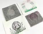 Two grey carved linoleum blocks and two printed sheets of paper, arranged on a flat surface. Each piece of paper is printed using one of the blocks. The foreground paper is printed in a dark gradient and depicts the hippo. The background paper is printed using pastel colors and shows trees, water texture, and some grass.
