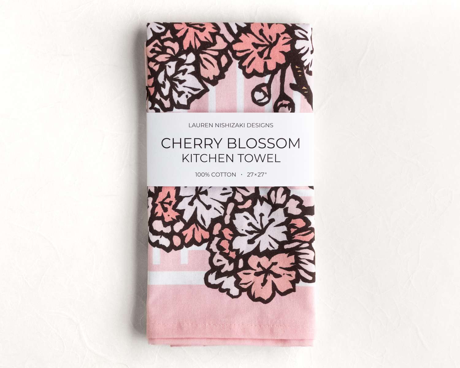A folded kitchen towel featuring a pink and black illustration of ginkgo leaves. A white belly band reads 'Lauren Nishizaki Designs; Cherry blossoms kitchen towel; 100% cotton; 27x27