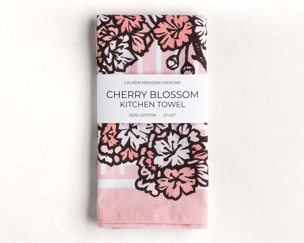 A folded kitchen towel featuring a pink and black illustration of ginkgo leaves. A white belly band reads 'Lauren Nishizaki Designs; Cherry blossoms kitchen towel; 100% cotton; 27x27