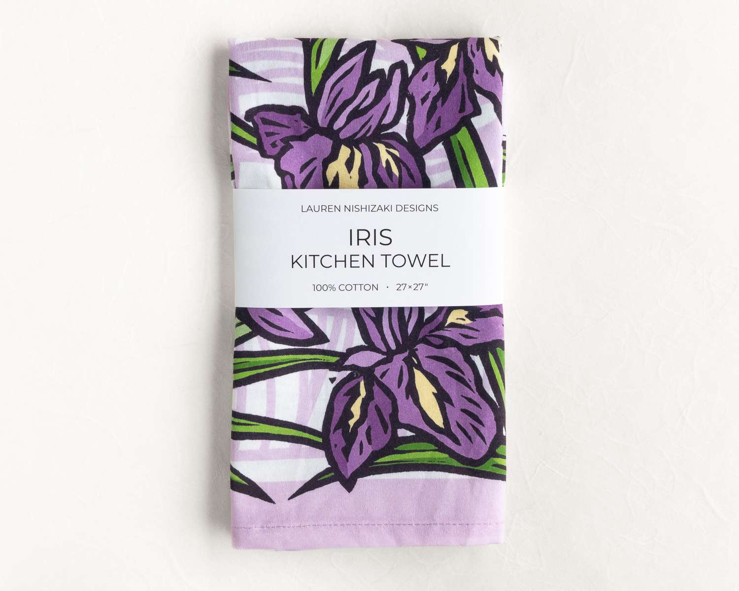 A folded kitchen towel featuring a purple and black illustration of irises and their leaves. A white belly band reads 'Lauren Nishizaki Designs; Iris kitchen towel; 100% cotton; 27x27
