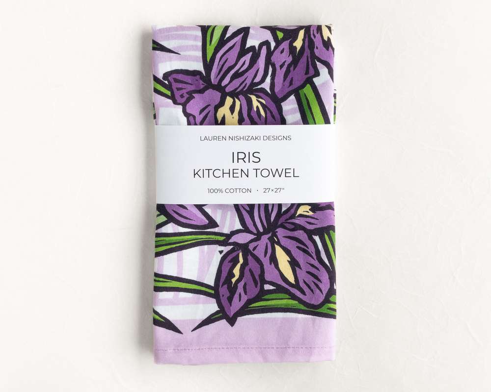 A folded kitchen towel featuring a purple and black illustration of irises and their leaves. A white belly band reads 'Lauren Nishizaki Designs; Iris kitchen towel; 100% cotton; 27x27