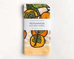 A folded kitchen towel featuring an orange and black illustration of persimmon with green leaves. A white belly band reads 'Lauren Nishizaki Designs; Persimmon kitchen towel; 100% cotton; 27x27