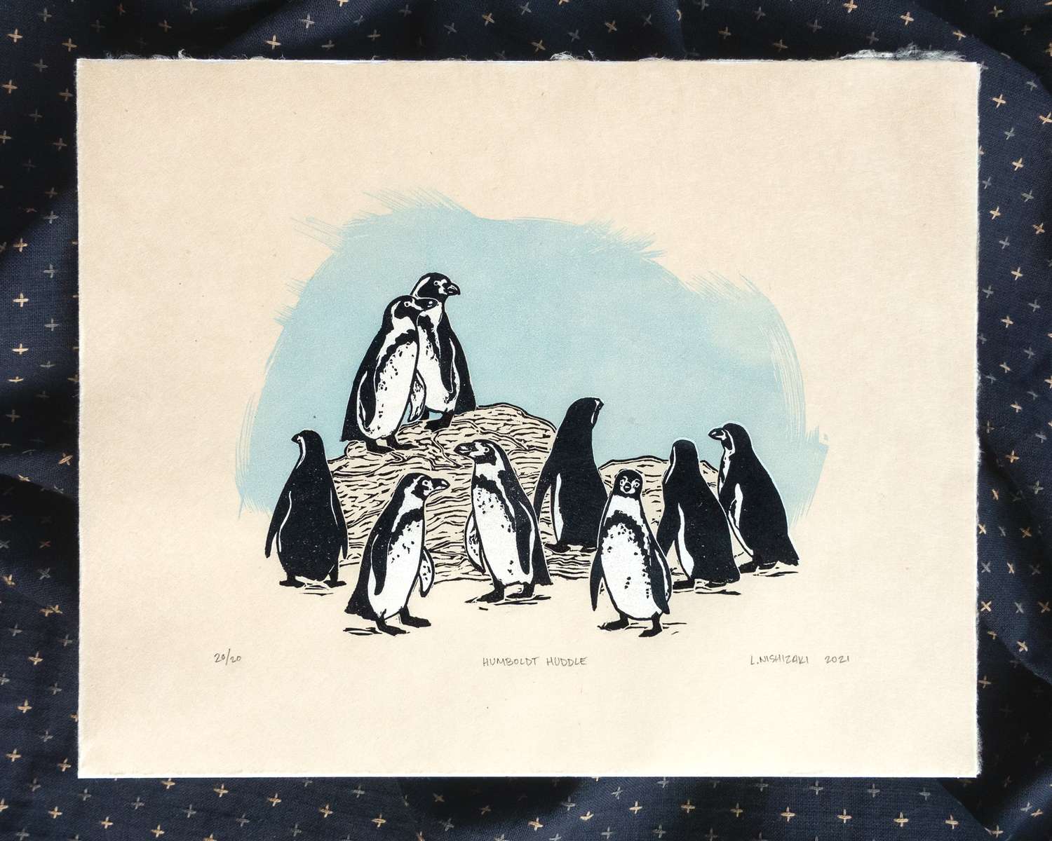 A brown rectangular print depicting 9 black and white Humboldt penguins (penguins with black-spotted bellies) standing at different heights on a rock. Behind them, the sky is colored blue and looks like its made from brushstrokes. The print sits on top of a rumpled piece of dark fabric.